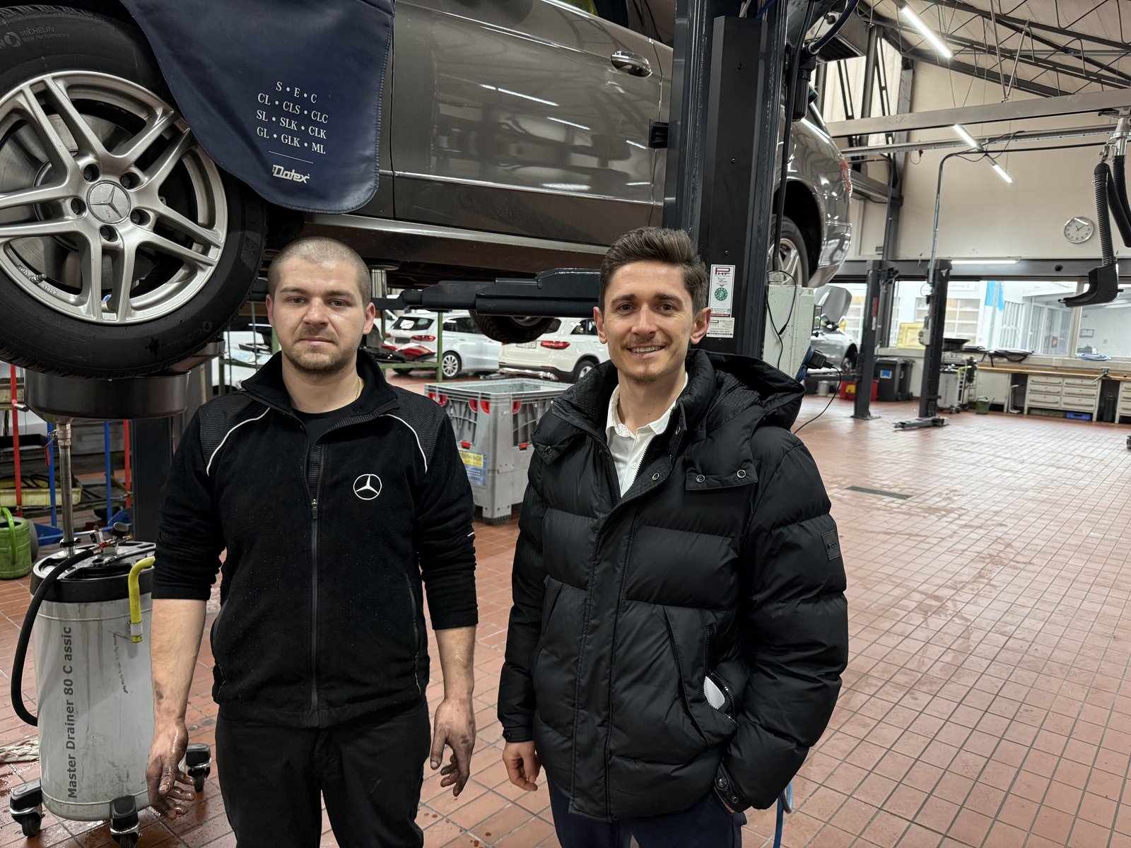 Kfz-Mechaniker aus dem Balkan an seinem neuen Arbeitsplatz in einer deutschen Markenwerkstatt, mit Personalvermittler unter dem Fahrzeug auf der Hebebühne